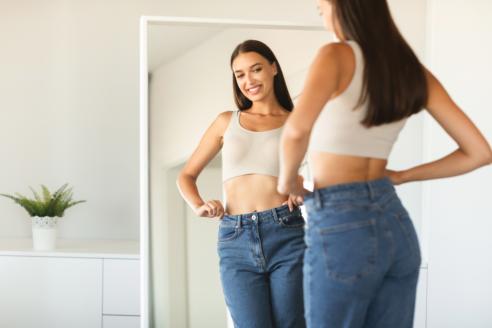 Cheerful young woman posing wearing jeans and smiling to her reflection in mirror after successful weight loss